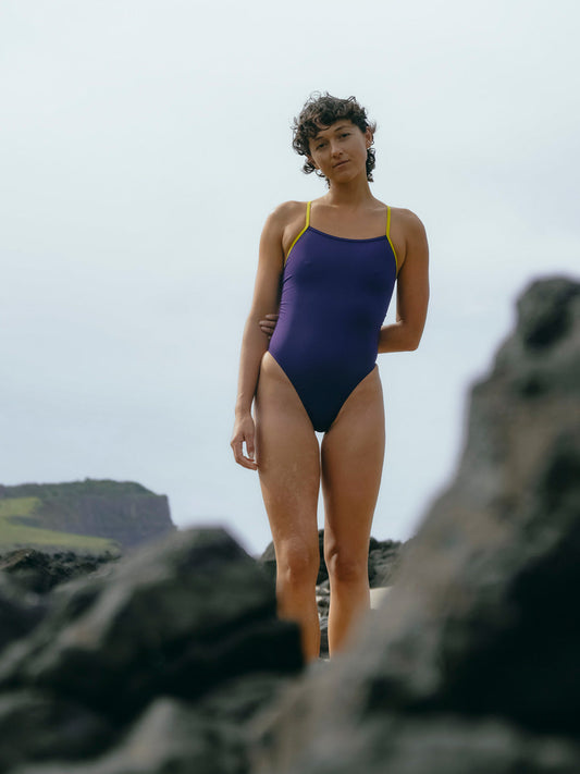 JAYD Swimwear model Sophie in a purple acai Lennox one piece swimsuit standing on rocky terrain with a natural landscape background