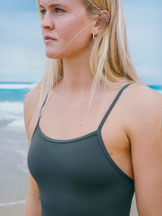 JAYD Swimwear model wearing the Albi one piece in carbon grey, staring out towards the ocean with waves in the background.