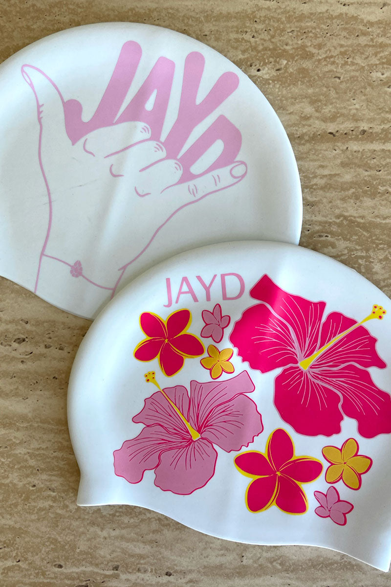 Pink JAYD swim caps Shaka and Hibiscus Haven on a stone background.