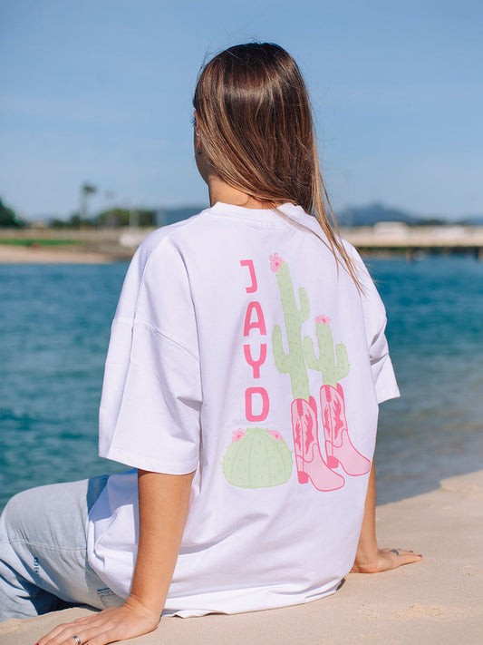 Back view of a model in JAYD Swimwear's white oversized tee, printed with a playful pink JAYD word mark, cactus, and cowgirl boots graphic.