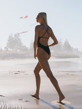Athlete in a cacao brown fixed back one piece walking on a beach with trees in the background.
