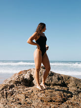 Woman in a black JAYD one piece standing on a rock at the beach with clear blue sky. She is wearing the Albi style