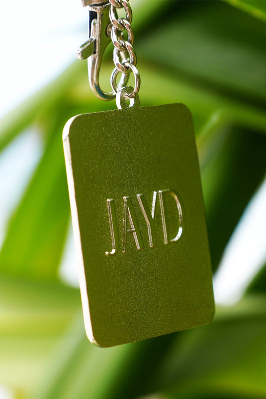 Back of JAYD Swimwear shell stamp keyring, featuring embossed JAYD lettering, hanging on a branch.
