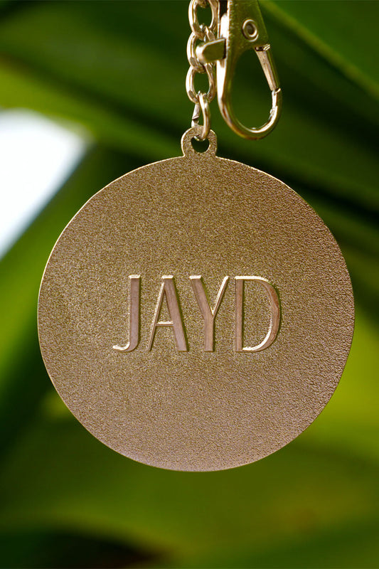 Back of JAYD Swimwear keychain with 'JAYD' engraved on a blurred green background