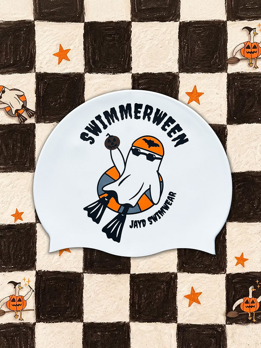 A white swim cap with a Halloween-themed graphic featuring a ghost swimmer in orange and black, with the text 'SWIMMERWEEN' and 'JAYD SWIMWEAR', displayed on a black and cream checkered background with small pumpkin and star illustrations.