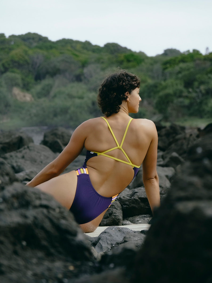 JAYD Swimwear model sitting on a rocky beach in the Lennox contrast one piece in Acai purple and daffodil yellow.