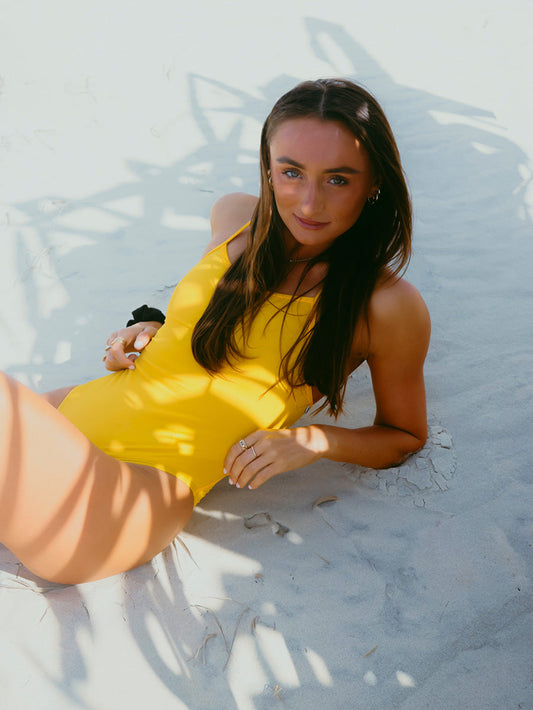 Triathlete Lauren laying on the sand at the beach in the JAYD Airlie one piece in Ignite yellow.