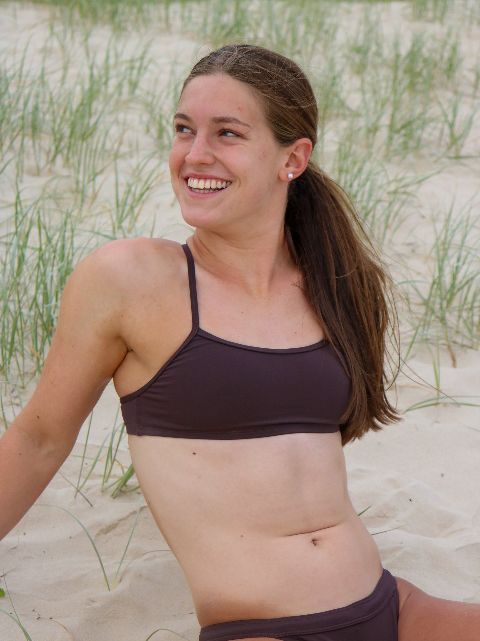 Front view of a female athlete in a JAYD Swimwear Torquay bikini top in cacao, sitting on a beach.