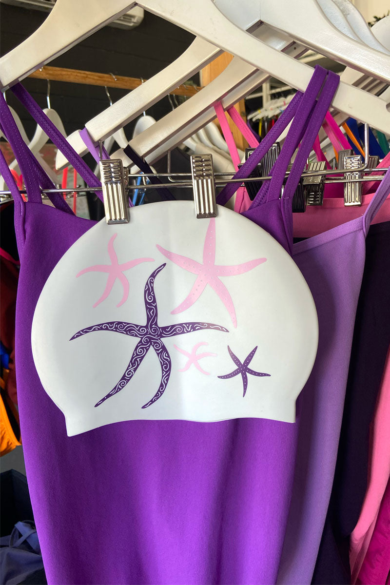 JAYD swim cap Starfish, hanging on a rack with purple and pink JAYD one piece swimsuits.