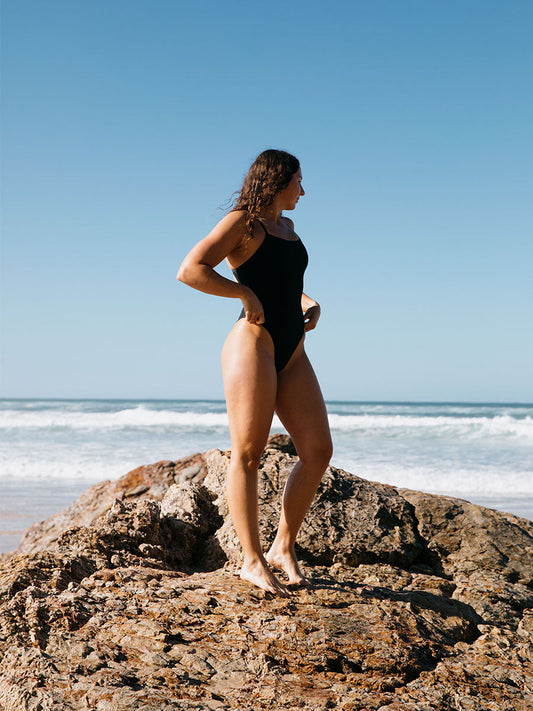 Woman in a black JAYD one piece standing on a rock at the beach with clear blue sky. She is wearing the Albi style