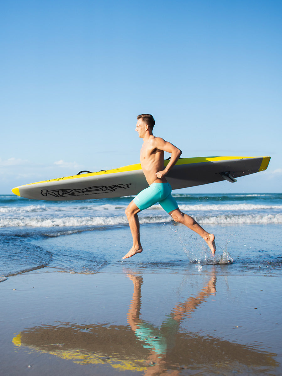A shirtless man running through shallow ocean water on a beach, carrying a yellow ski under his arm. He is wearing turquoise FINIS Swim Jammers, with the ocean, waves, and blue sky in the background.