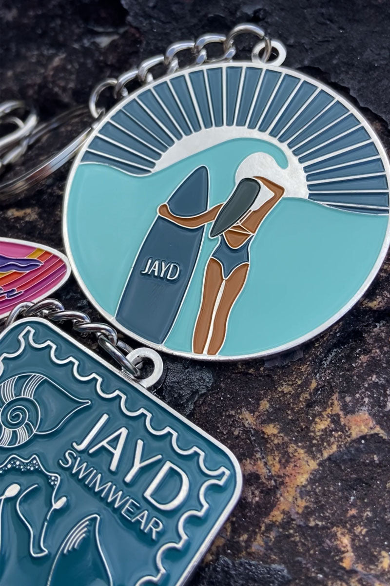 JAYD Swimwear keychain featuring a woman with a surfboard staring into the distant sunset on a beach, sitting on a rock with other colourful keychain designs.