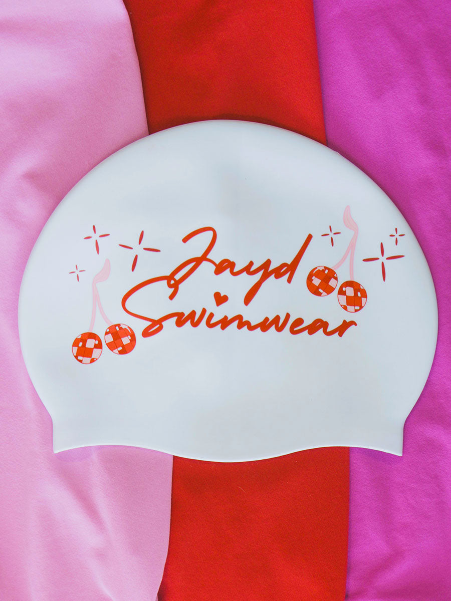 White swim cap with cursive JAYD Swimwear text and red and pink cherry disco balls.
