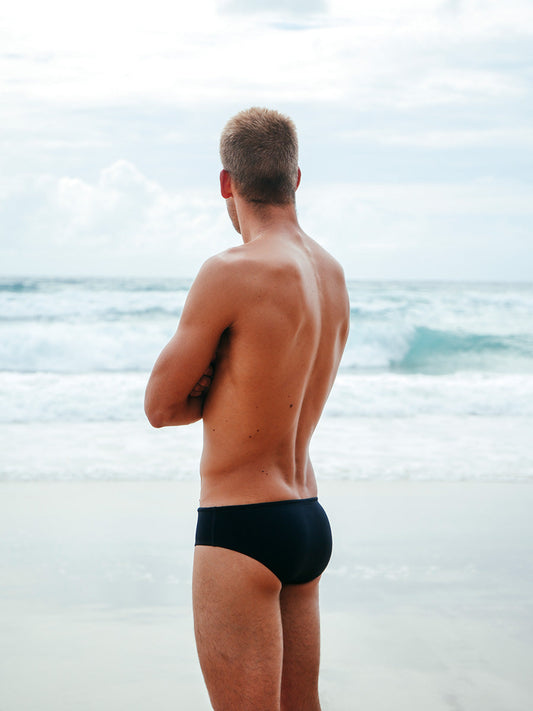 JAYD Swimwear model wearing the black Ramshark men's swim bottoms, showcasing the comfortable moderate coverage fit, staring off into the distance at the beach.