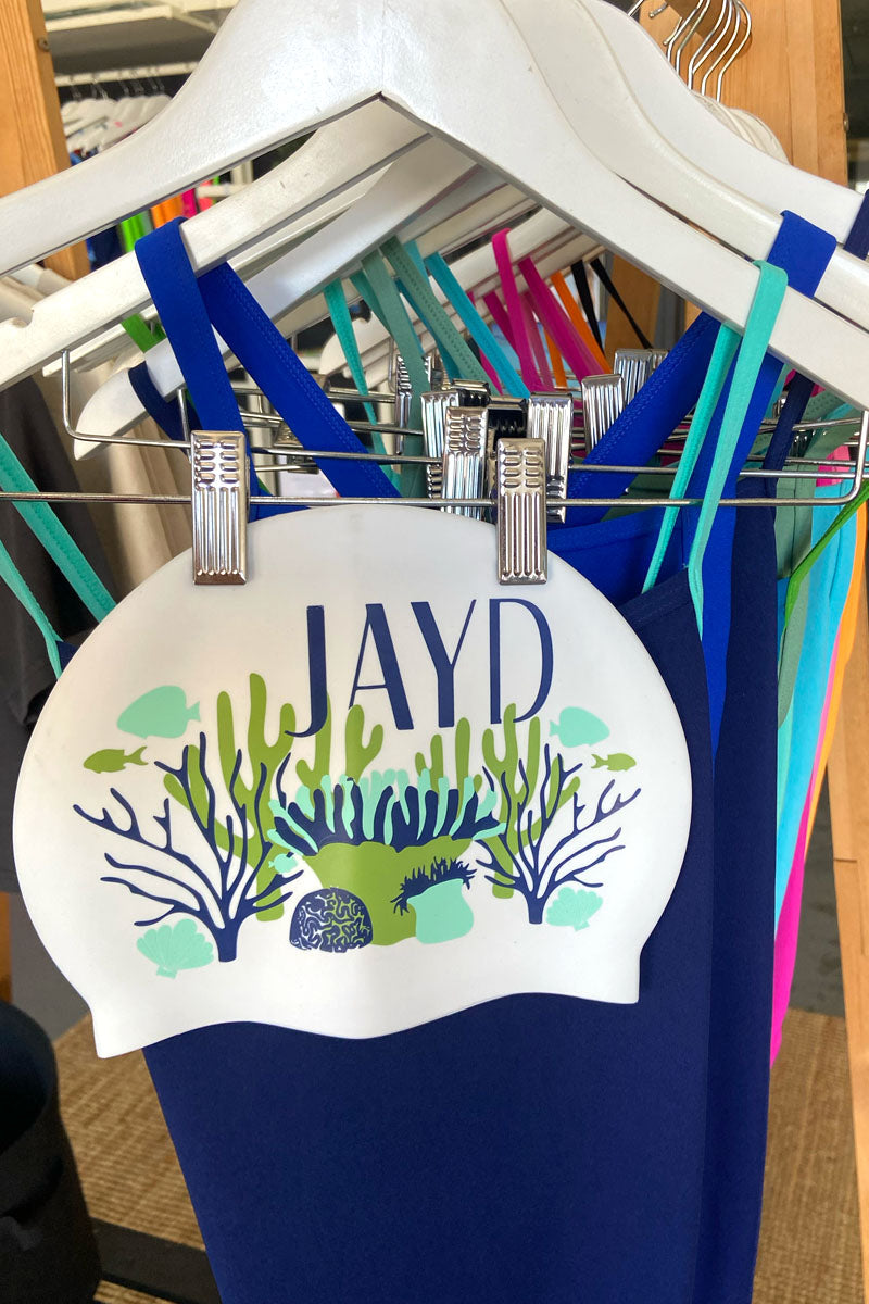 White swim cap design featuring JAYD logo & blue and green coral illustrations, paired with JAYD one piece blue swimsuits.