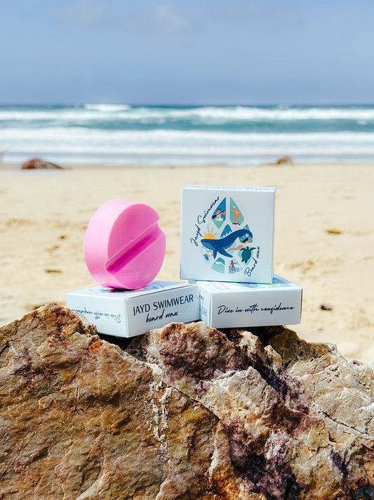 JAYD Swimwear pink circular surf wax arranged in a stack on top of a rock with the beach and ocean in the distance.