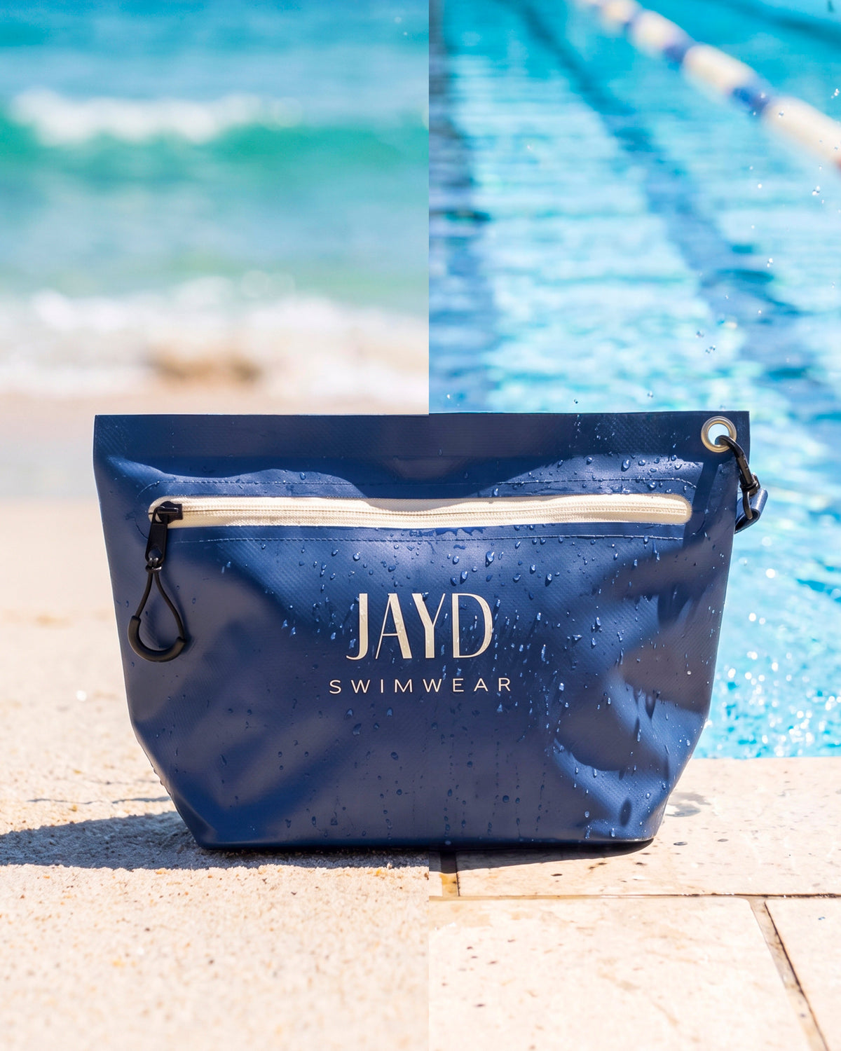 Blue JAYD Swimwear dry bag sitting on the edge of a pool with water droplets running down the waterproof material.