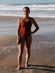 Woman in a Tallow swimsuit by JAYD Swimwear, standing on a beach with ocean waves in the background